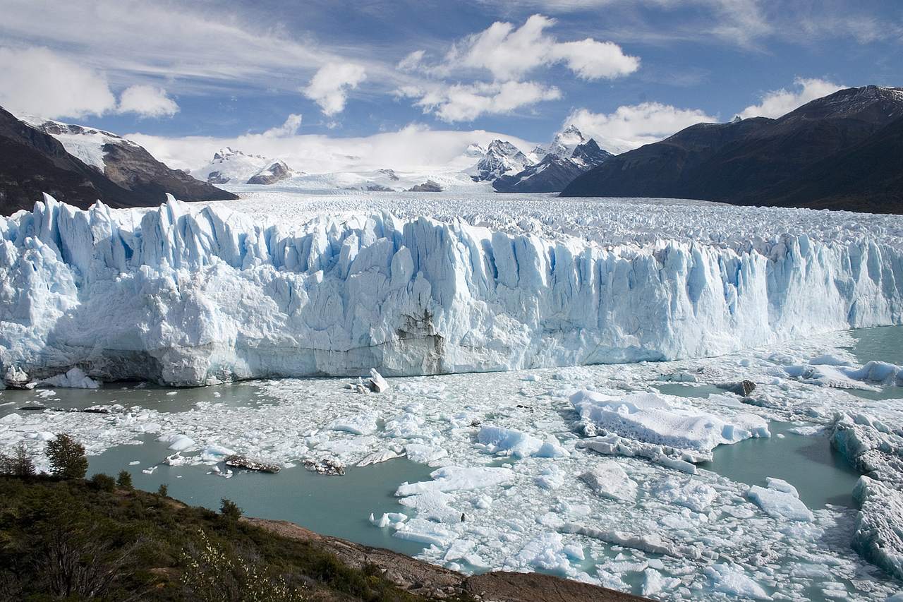 Perito Moreno-glaciärens vita och blå isvägg reser sig över en vik full av isflak, med snöklädda berg och moln i bakgrunden