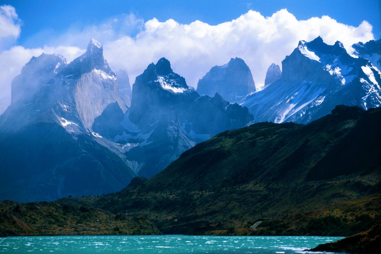Paine-massivets taggiga granittoppar med snö och moln reser sig över gröna kullar och en turkos sjö i Torres del Paine