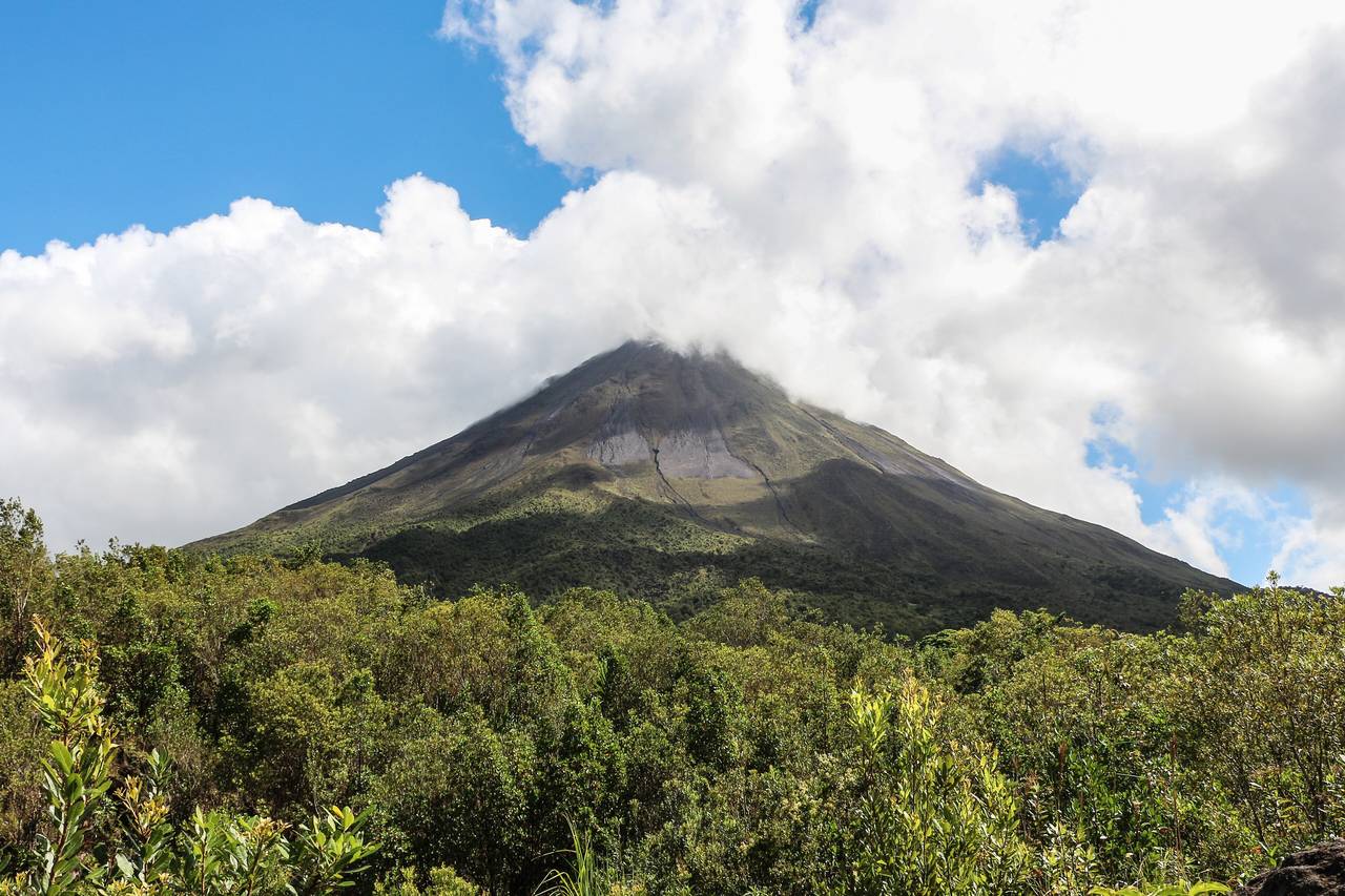 Den kägelformade vulkanen Arenal reser sig över tät grön regnskog under blå himmel med vita moln