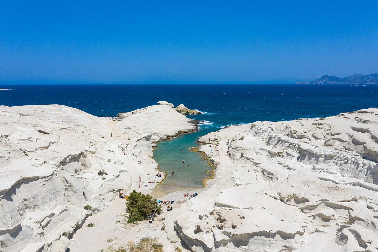 Flygbild över Sarakiniko på Milos med vita, vågformade vulkanklippor, en smal turkos vik och mörkblått hav bakom