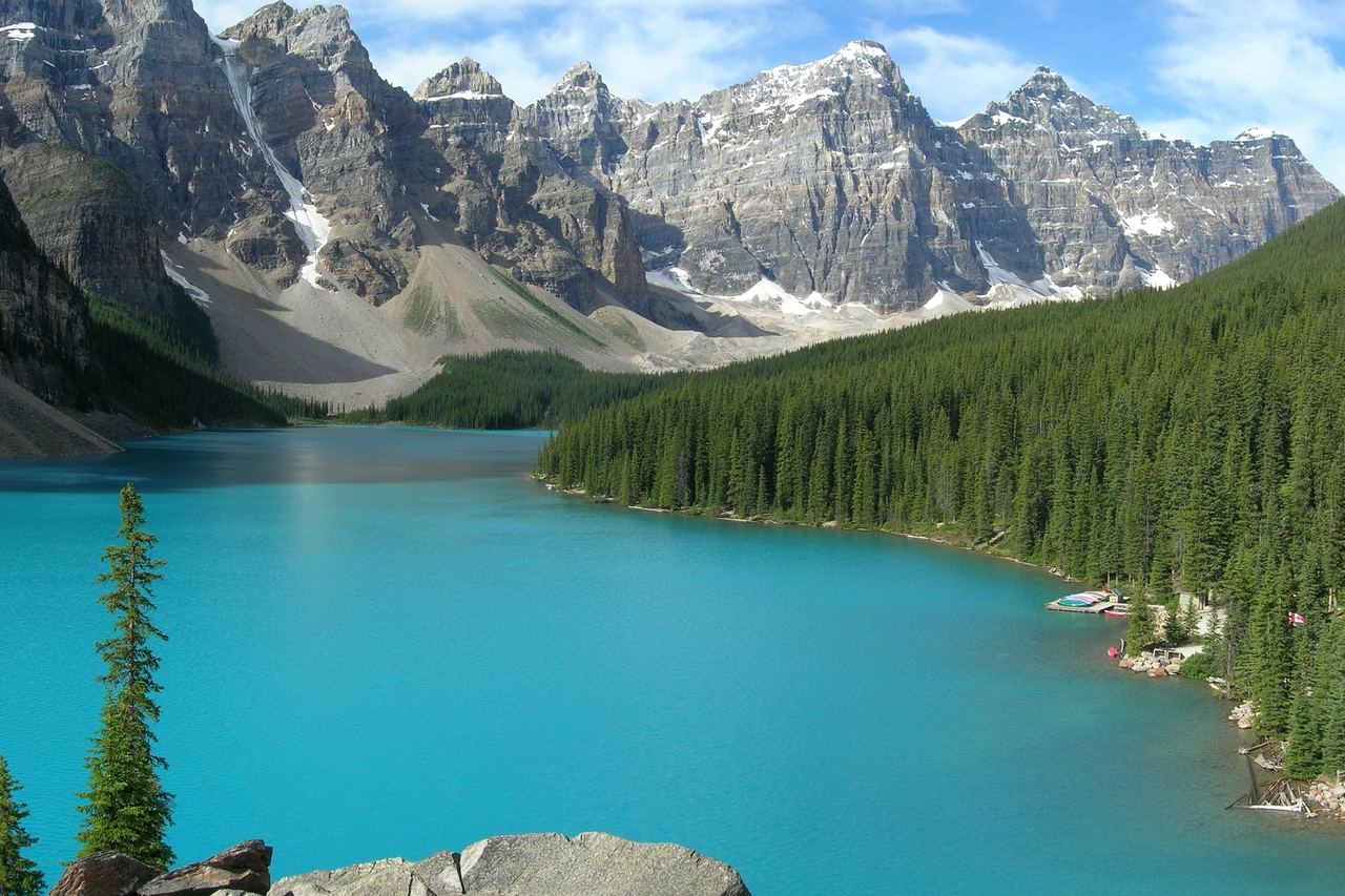 Den turkosa Moraine Lake omgiven av granskog, med en rad branta, snöfläckiga bergstoppar i Valley of the Ten Peaks under en blå himmel