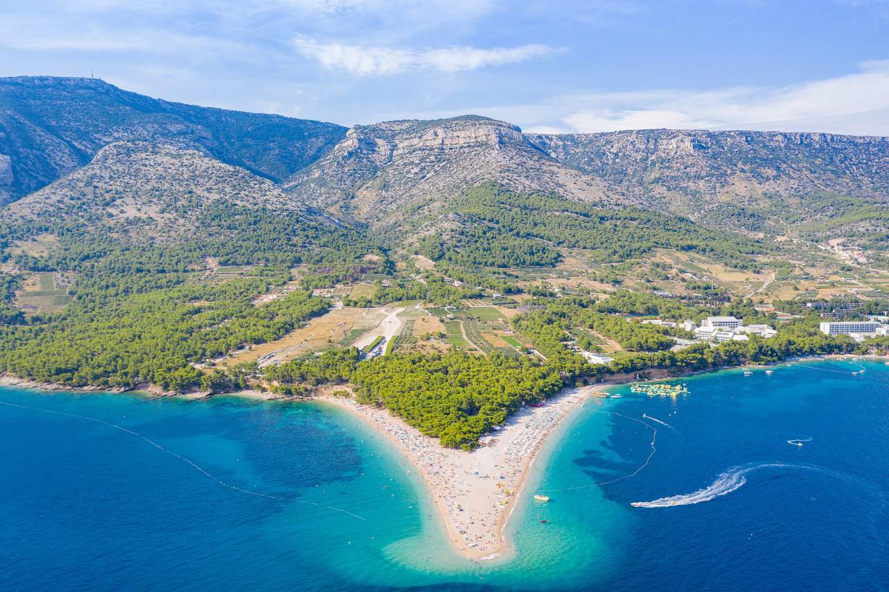 Flygbild över stranden Zlatni Rat vid Bol på Brač, en lång udde av ljus sten och sand som sticker ut i turkost vatten, med tallskog och kala berg bakom