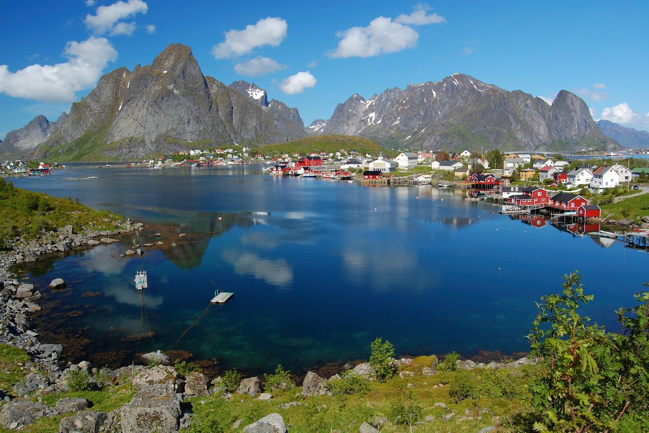 Fiskeläget Reine i Lofoten med röda och vita trähus längs en blank fjord, omgivet av spetsiga, branta berg under en blå himmel med vita moln