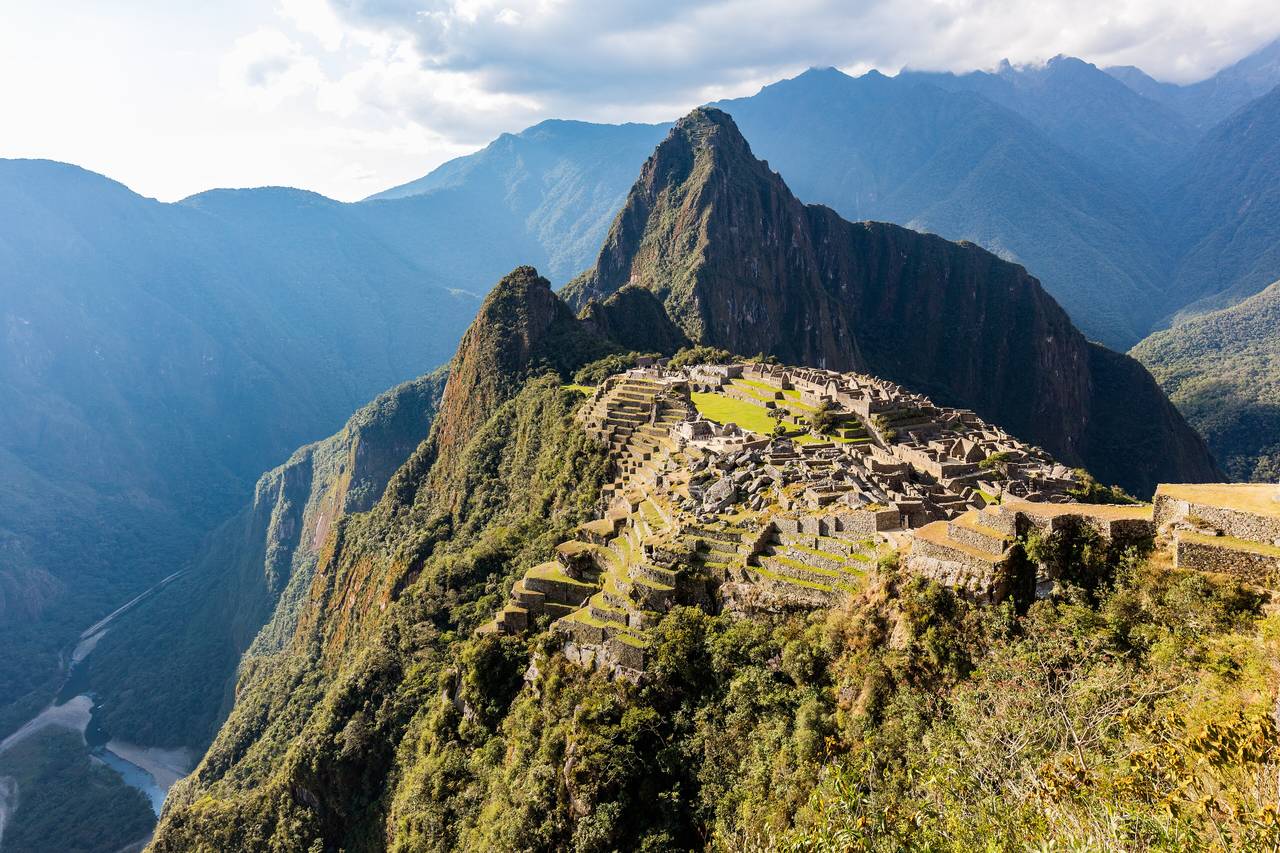 Inkastaden Machu Picchu på en grön bergsrygg med det spetsiga berget Huayna Picchu bakom och djupa dalar runt omkring i eftermiddagsljus