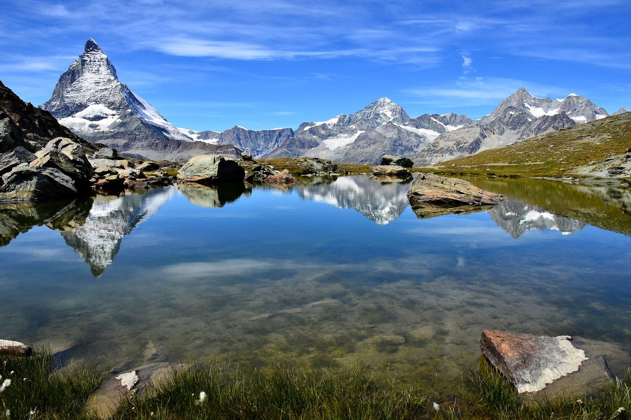 Matterhorns spetsiga, snöfläckiga topp och en rad vita alptoppar speglar sig i den stilla Riffelsee, med stenar och gräs i förgrunden under en blå himmel
