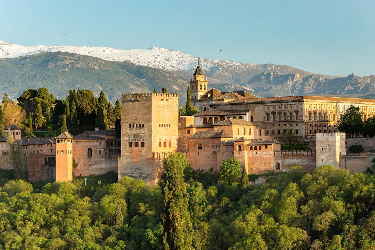 Alhambra i Granada i varmt kvällsljus, med borgens torn och murar ovanför grönskande träd och snöklädda Sierra Nevada i bakgrunden