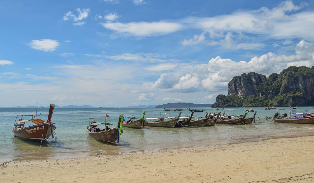 Långsvansbåtar förtöjda vid den ljusa sandstranden Railay West i Krabi, med turkost vatten och en kalkstensklippa i bakgrunden