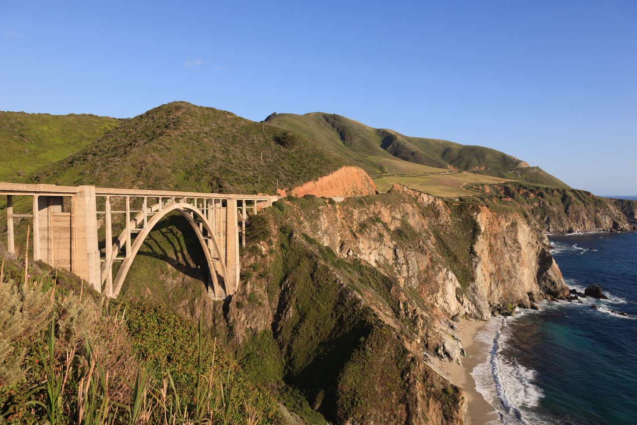 Bixby Creek Bridge, en ljus betongbro med en hög båge, spänner över en ravin vid Big Surs gröna kustberg, med det blå Stilla havet nedanför