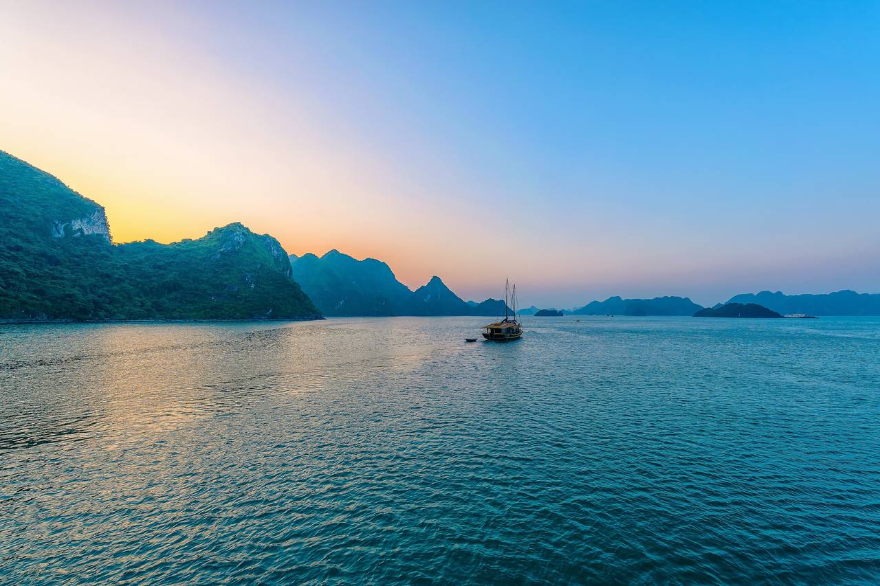 Solnedgång över Ha Long Bay, med kalkstensöar i silhuett, en ensam båt och stilla, turkost vatten under en himmel i orange och blått