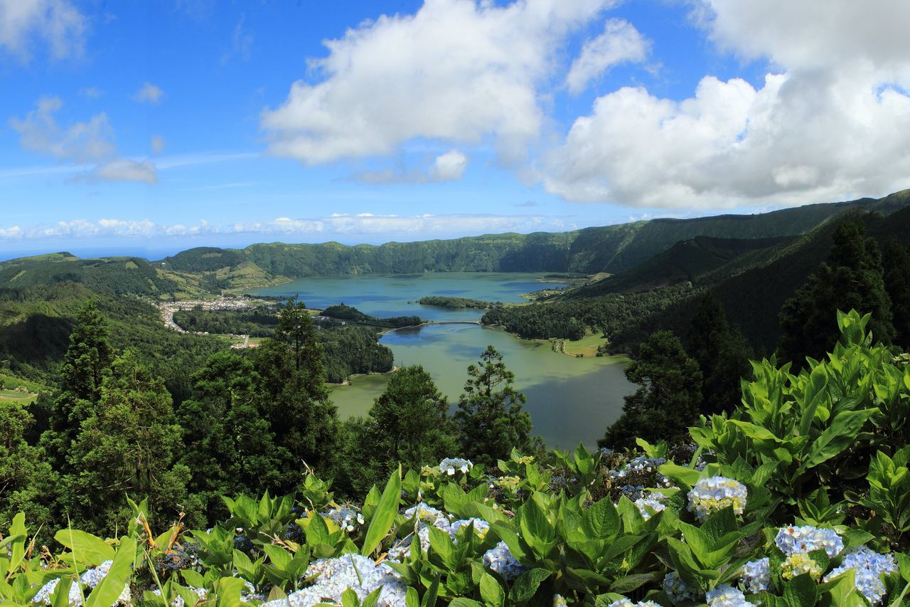 Kratersjöarna vid Sete Cidades på São Miguel sedda över blommande blå hortensior, med gröna kraterväggar och vita moln