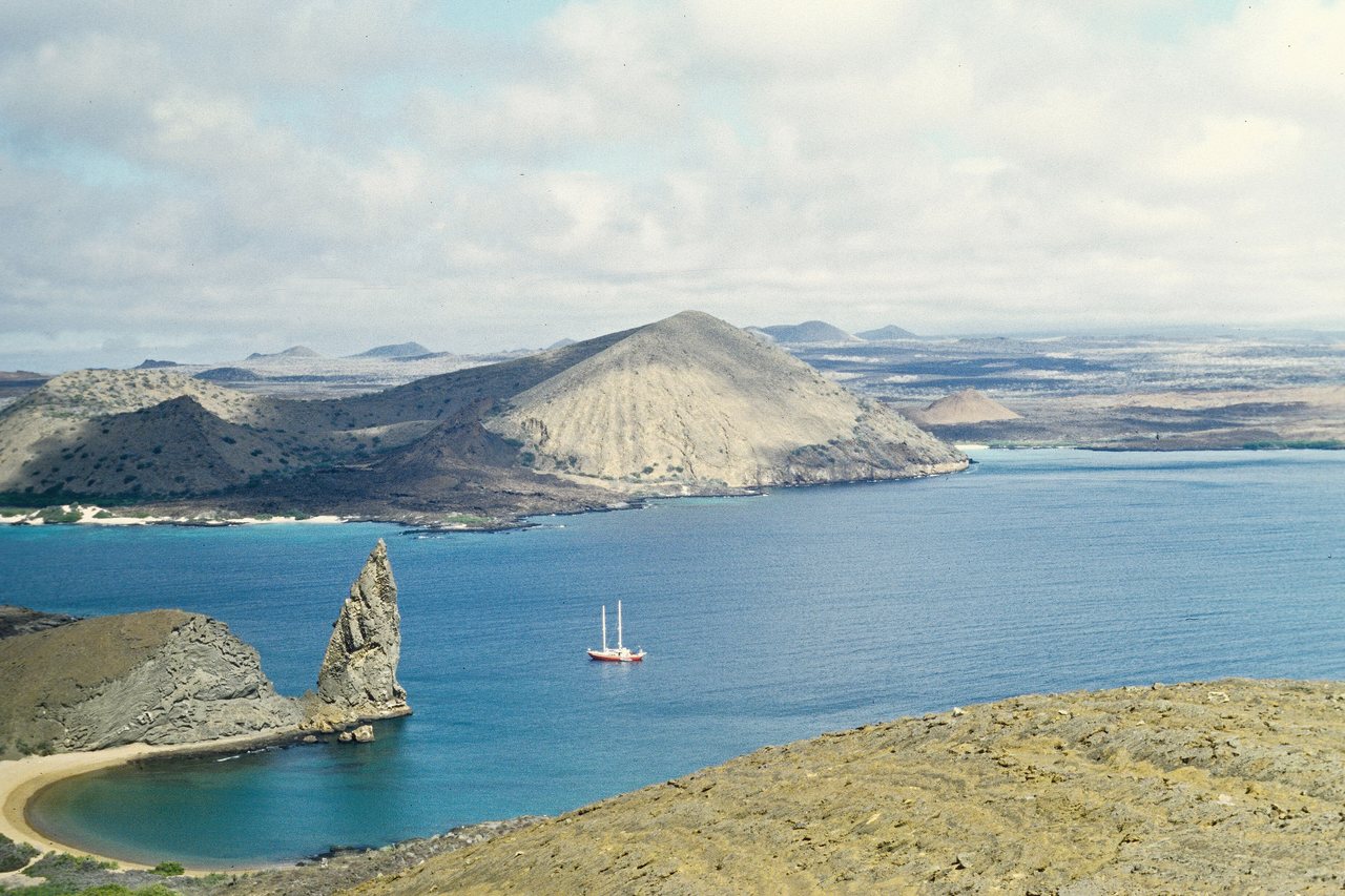 Utsikt från Bartolomé över den spetsiga klippan Pinnacle Rock och en ljus sandstrand i en turkos vik, med en segelbåt för ankar och vulkaniska öar i bakgrunden
