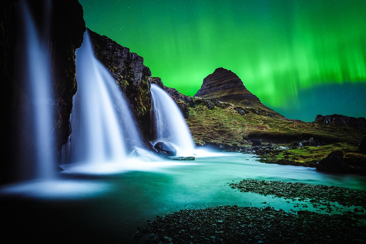 Grönt norrsken över det spetsiga berget Kirkjufell på Snæfellsnes, med vattenfallet Kirkjufellsfoss i förgrunden under en stjärnklar natthimmel