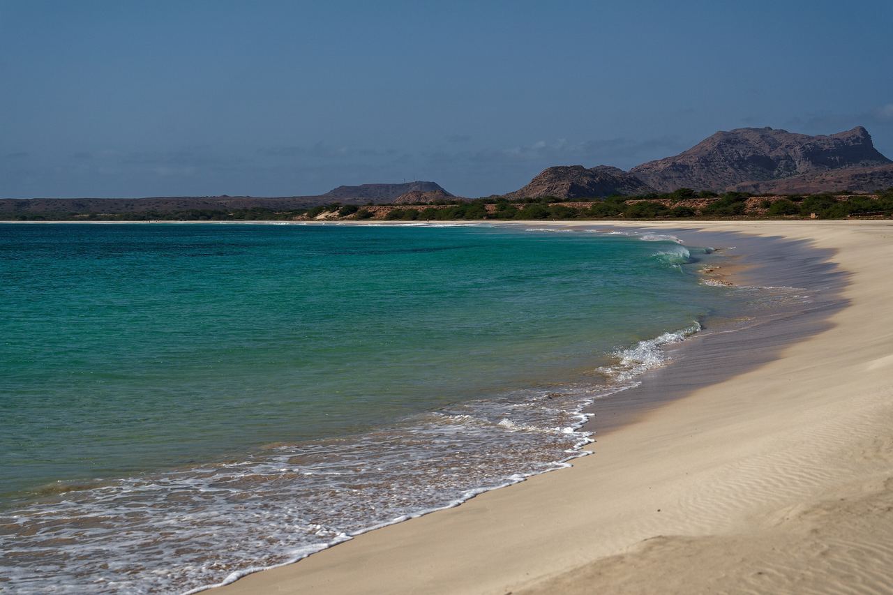 Turkost vatten och en lång, ljus sandstrand på södra Boa Vista i Kap Verde, med bruna berg vid horisonten