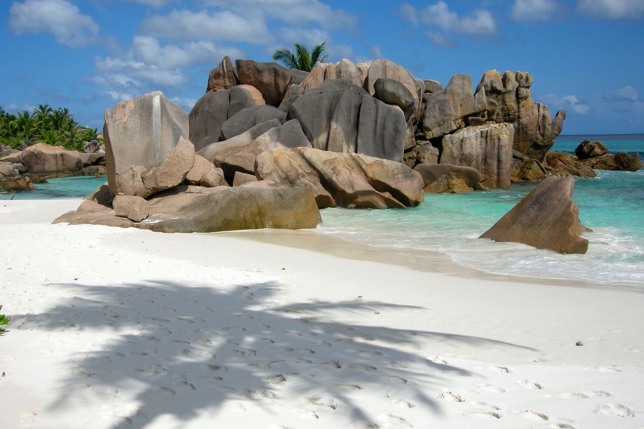 Stora rundade granitklippor i grått och rosa vid en strand med kritvit sand och klart turkost vatten på La Digue, med skuggan av en palm i förgrunden