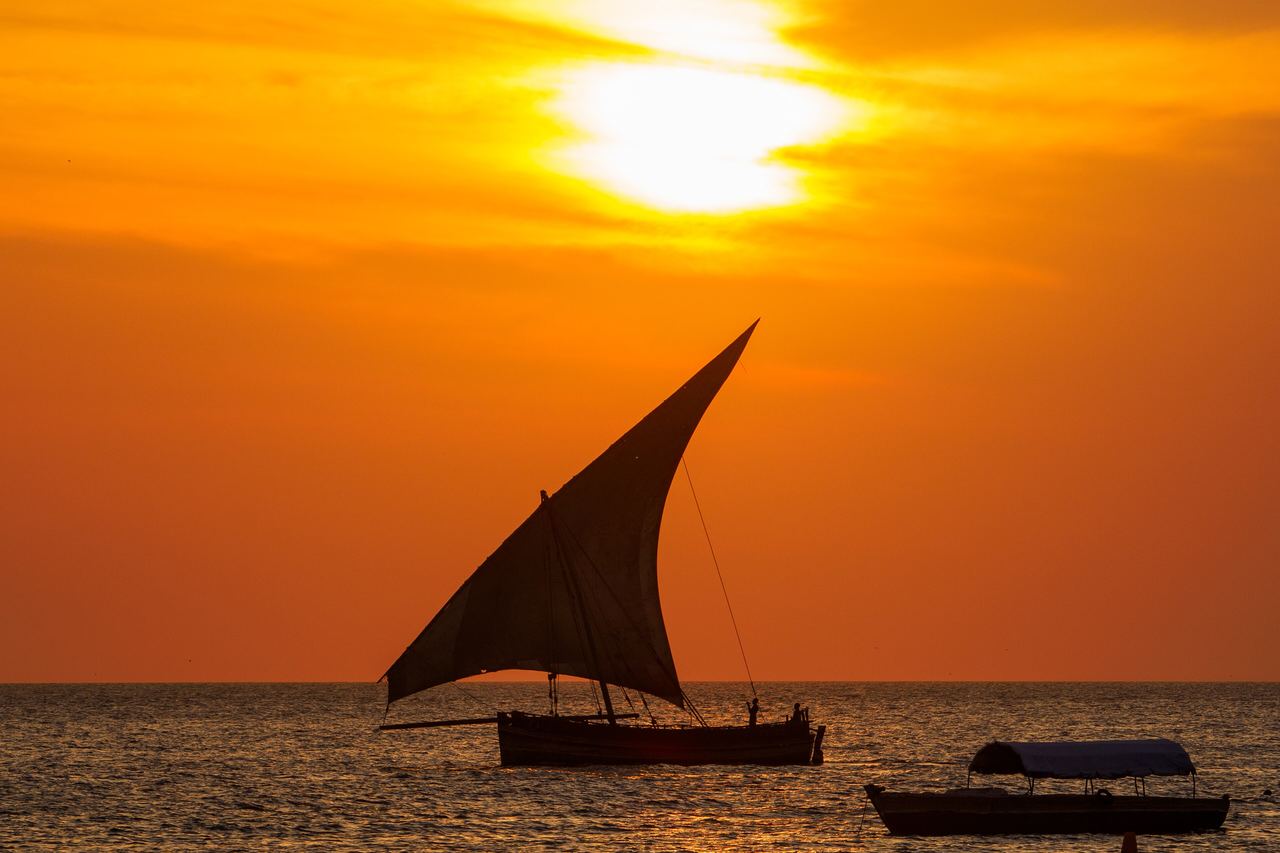 En dhow med triangulärt segel glider fram i siluett mot en glödande orange solnedgång över havet utanför Stone Town
